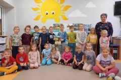 Grupa dzieci siedzi na podłodze, za nimi toją. W tle widac ścianę z namalowanym słoneczkiem.