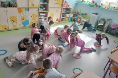 Na szarej podłodze leżą kolorowe obręcze. Pomiędzy nimi na czworakach chodzą dzieci.