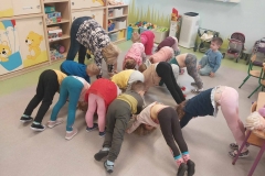 Grupa dzieci w kolorowych ubraniach i pani w czarnych spodniach robią "koci grzbiet"- dłonie i stopy oparte na szarej podłodze, kręgosłup wygięty w pałąk. Wzdłuż ściany ustawiona beżowa meblościanka.