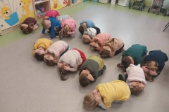 Dzieci w kuckach siedzą na szarej podłodze- nogi podkurczone, głowa schowana, ręce ułożone na karku. Pod ścianą ustawione pojemniki z klockami i 2 wózki dla lalek.