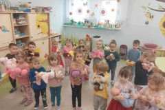 Dzieci w kolorowych ubraniach przytulają do siebie maskotki piesków, kotków, misiów i lalki. Obok ściany beżowa meblościanka.  W tle okno - na firanie zawieszone kolorowe kwiaty. Na parapecie- kwiaty, biedronki, pisanki, świąteczne kartki.