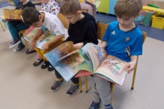 W sali przedszkolnej na krzesełkach ustawionych obok siebie siedzi sześcioro dzieci. Ich głowy są pochylone. Każde z nich przegląda książkę  z serii o Kubusiu Puchatku.   W tle widoczne szafki przedszkolne.