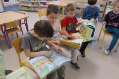 W sali przedszkolnej na krzesełkach ustawionych w półkolu  siedzi pięcioro dzieci. Mają pochylone głowy. Przeglądają książki  z serii o Kubusiu Puchatku.   W tle widoczne stoliki i szafki przedszkolne.