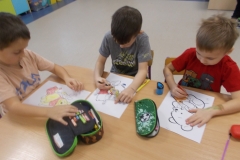 Przy stoliku siedzą trzej chłopcy. Mają na sobie czerwone, szare i kremowe koszulki z nadrukami. Kolorują obrazki konturowe z sylwetami Kubusia Puchatka. Na stole leża piórniki i temperówki. W tle widać szafki przedszkolne.