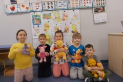 Na tle tablicy z obrazkami o Kubusiu Puchatku klęczy czworo dzieci: dwie dziewczynki i dwóch chłopców. Obok nich siedzi chłopiec w siadzie skrzyżnym. Dzieci trzymają w rękach maskotki: Kubusia Puchatka ( 2 dzieci), Tygryska, Prosiaczka oraz Kłapouchego.