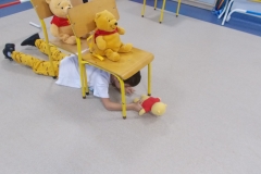 W sali przedszkolnej ustawione zostały dwa krzesełka, jedno za drugim. Na każdym siedzi pluszowy miś - Kubuś Puchatek. Pod krzesłami czołga się chłopiec w białym podkoszulku i żółtych spodniach. Przed sobą trzyma misia, też Kubusia Puchatka. Po prawej stronie widać płotki gimnastyczne, obok krzesełko.