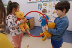 Chłopiec ubrany na niebiesko oraz dziewczynka w biało - różowym stroju trzymają w rękach pluszowe misie - Kubusie Puchatki oraz czerwoną plastikową obręcz. Za obręczą stoi chłopiec w niebieskiej bluzie i przerzuca maskotkę Kłapouchego przez obręcz. Z przodu widoczne dłonie dziecka w żółtej bluzce. Ma ono złapać zabawkę.  W tle tablica z pokolorowanymi obrazkami misia.