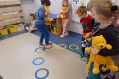 Grupa dzieci trzymając w rękach pluszowe zabawki przemieszcza się po plastikowych obręczach ułożonych na podłodze w sali przedszkolnej. W tle widać chłopca siedzącego po turecku oraz stojącą dziewczynkę. Trzyma ona żółtego misia. Po lewej stronie pod oknem widać pudełka z klockami.