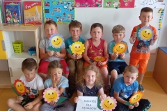 Na ławce przed tablicą oraz na podłodze przednią siedzi ośmioro dzieci, jedno stoi z prawej strony. Ubrane są w kolorowe stroje. Każde z nich trzyma w ręce kolorowy kwiat, który w swoim środku ma wyklejoną uśmiechniętą buzię. Dziewczynka siedząca w środku trzyma dodatkowo białą kartkę z napisem: Kwiatek dla sąsiadów. W tle na tablicy przypięte są kolorowe kartki z mapami mentalnymi. Z lewej strony stoi szafka z książkami.