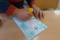 Częściowo widoczne ręce dziecka. W jednej z nich trzyma ono klej i smaruje nim kartonik do przyklejenia na pokolorowanej na zielono kartce. Odrysowana jest na niej dłoń a na palcach wyrysowane postacie członków rodziny.