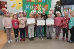 Na zielonym płótnie napis: "Dzień Puzzli". Przed nim na szarej podłodze stoi grupa dzieci z opaskami puzzli na głowie. 2 chłopców i 1 dziewczynka trzymają w rękach dyplomy.