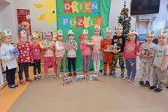 Grupa 13 dzieci w kolorowych strojach i opaskach na głowie stoi w półkolu. W rękach trzymają 6 pudełek puzzli. Na szarej podłodze leżą 3 pudełka z  puzzlami. Za nimi na zielonym tle napis " Dzień Puzzli".