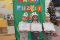 Troje dzieci stoi na tle dekoracji. W rękach trzymają dyplomy z napisem: " Mistrz w układaniu puzzli". W tle kolorowa choinka.