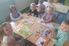 .Sala przedszkolna . Grupa dzieci ubrana na kolorowo siedzi przy stoliku. Dzieci układają puzzle drewnine różnego formatu i puzzle książeczkowe. Przy drugim stoliku siedzą dwie dziewczynki odwrócone tyłem.