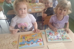 Sala przedszkolna . Na pierwszym planie dwie dziewczynki ubrane na kolorowo układają puzzle. Jedna z nich układa puzzle o tematyce zimowej – (Zabawy na śniegu ) druga o tematyce letniej – (Na plaży ). Za dziewczynkami widać fragmenty stolików przy których również siedzą dzieci . Widać rozłożone na stolikach puzzle.