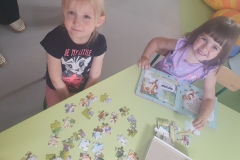 Trójka dzieci; dziewczynka i dwóch chłopców układają na podłodze puzzle dużego formatu .Przed dziećmi leżą pudełka po puzzlach. Dzieci się uśmiechają.