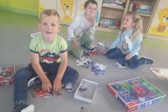Trójka dzieci; dziewczynka i dwóch chłopców układają na podłodze puzzle dużego formatu .Przed dziećmi leżą pudełka po puzzlach. Dzieci się uśmiechają.