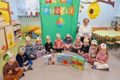 Grupa dzieci w kolorowych strojach siedzi na podłodze tworząc półkole. Przed nimi  stoją 3 pudełka  z puzzlami. Z tyłu na zielonym tle widnieje napis Dzień  Puzzli