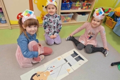 Trzy dziewczynki siedzą na podłodze.  Przed nimi na podłodze leży ułożona z puzzli postać lekarza. W tle szafki