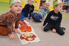 Maria Wilkosz: Czterech chłopców siedzi na podłodze,  przed nimi z ułożonych puzzli leży postać  strażaka.  W tle kolorowe szafki. 