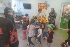 Sala przedszkolna . Dzieci ubrane na kolorowo z opaskami kwiatów na głowach stoją w kole i poruszają się po obwodzie koła w rytm muzyki. Zabawę obserwują rodzice.