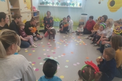 Rodzice z dziećmi w kole siedzą na podłodze . W środku koła na podłodze leżą rozrzucone papierowe sylwety kolorowych kwiatów.