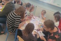 Sala przedszkolna . Rodzice z dziećmi przy stolikach wykonują pracę plastyczną pt; „ Kolorowa łąka”- wycinają motylki, kwiatki, biedronki, itp. . Na stolikach rozlożone materiały papiernicze , kleje , bibuła, kolorowe kartki i kredki