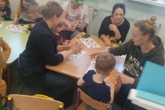 Sala przedszkolna . Rodzice z dziećmi przy stolikach wykonują pracę plastyczną pt; „ Kolorowa łąka”- wycinają motylki, kwiatki, biedronki, itp. . Na stolikach rozlożone materiały papiernicze , kleje , bibuła, kolorowe kartki i kredki