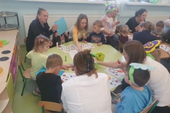 Sala przedszkolna . Rodzice z dziećmi przy stolikach wykonują pracę plastyczną pt; „ Kolorowa łąka”- wycinają motylki, kwiatki, biedronki, itp. . Na stolikach rozlożone materiały papiernicze , kleje , bibuła, kolorowe kartki i kredki