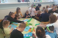 Sala przedszkolna . Rodzice z dziećmi przy stolikach wykonują pracę plastyczną pt; „ Kolorowa łąka”- wycinają motylki, kwiatki, biedronki, itp. . Na stolikach rozlożone materiały papiernicze , kleje , bibuła, kolorowe kartki i kredki