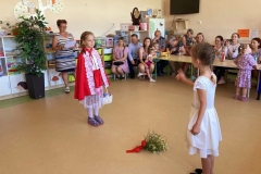 Czerwony Kapturek i mama rozmawiają ze sobą. Między nimi na podłodze polne kwiatki. W tle rodzice.