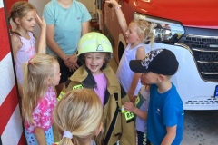 Kilkoro dzieci stoi przed wozem strażackim, jedna dziewczynka ubiera się w mundur strażaka.