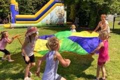 Kilkoro dzieci z wychowawczynią bawią się chustą animacyjną. W tle kolorowy dmuchaniec.