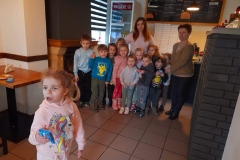  Dzieci stoją w grupie. Za nimi właścicielka pizzerii i wychowawczyni grupy. Przed nimi stoi dziewczynka w różowej bluzie.