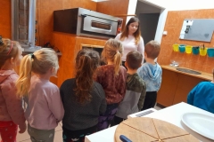 Dzieci stoją przy piecu, czekają, aż pizza upiecze się. Pani stoi koło pieca, opowiada dzieciom jak się pizza piecze.