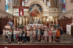 Grupa dzieci w kolorowych strojach stoi przed ołtarzem ozdobionym kolorowymi obrazami i kwiatami. Kilka dziewczynek ma na głowie czapki. Obok dzieci stoi ksiądz i 2 panie. Za nimi duży, brązowy krzyż. Przed nimi na posadzce leży bordowy chodnik.