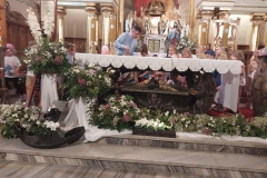 Pod głównym ołtarzem sarkofag bł. Karoliny. Obok niego dużo białych i różowych kwiatów. Kilka dzieci klęczy, a kilka stoi przy sarkofagu. Nad nimi pochyla się ksiądz w niebieskiej koszuli.
