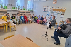 W sali przedszkolnej, na krzesełkach ustawionych w półkolu siedzą dzieci. Mają na sobie kolorowe stroje. Naprzeciwko nich siedzi mężczyzna grający na harmonii. Przed nim stoi statyw z nutami. W głębi przy ścianie siedzi nauczycielka. Na oknach widać zimowo - świąteczne dekoracje. W tle tablice z planszami liter i cyfr i zimowymi obrazkami.