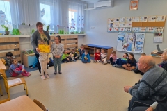 Na podłodze w sali przedszkolnej siedzą dzieci. Naprzeciw nich widać grupę starszych osób - seniorów. Dwie z dziewczynek stoją zwrócone twarzą w ich kierunku. Jedna z nich trzyma żółtą kartę ozdobioną kwiatami i z nieczytelnym napisem. Za dziewczynkami stoi nauczycielka. Po lewej stronie widać stoliki i krzesełka.