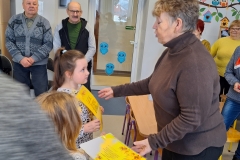 Dwie dziewczynki wręczają kobiecie żółte ozdobne karty z nieczytelnymi napisami. Kobieta jest w starszym wieku. Ubrana jest w szary golf i ciemne spodnie. Z prawej strony za nia widać częściowo trzy kobiety. Za nimi widać ścianę pomalowaną           w motywy dekoracyjne z sowami. Z lewej stoją dwaj starsi mężczyźni. Obok nich widać drzwi wejściowe do sali.