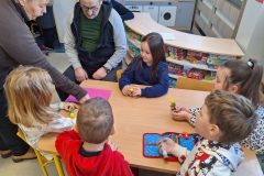 Przy stoliku przedszkolnym siedzi pięcioro dzieci. Trzymają w rękach kleje. Na stoliku leży czerwono niebieski piórnik i różowa kartka. Stojąca z lewej strony kobieta pokazuje dzieciom jak zagiąć kartkę. Obok na krzesełku siedzi mężczyzna w rozpiętej niebiesko - granatowej bluzie. W tle szafki przedszkolne.