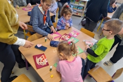 Przy stoliku w sali przedszkolnej widać dwie siedzące dziewczynki i stojącego chłopca. Jedna z dziewczynek ubrana jest w niebieską bluzę we wzory, druga  w różową. Chłopiec ma bluzę seledynową. Przy nich siedzi kobieta w niebieskiej bluzie i czerwono - białej apaszce. Nosi okulary. Obok stoi druga kobieta ubrana  w żółtą bluzę i ciemne spodnie. Na stoliku rozłożone są kolorowe  kartki z bloku złożone na pół. Na nich wyklejone są filiżanki na spodkach a na nich przyklejone drobne elementy dekoracyjne. Na stoliku leżą piórniki i kleje. W tle częściowo widoczne inne dzieci i dorosłe osoby.