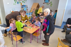 Przy stolikach przedszkolnych siedzą i stoją dzieci i osoby dorosłe. Tworzą wyklejanki na kolorowych kartkach. Pośrodku widać stojącą starszą kobietę. Ubrana jest w ciemny sweter z różowo - niebieskim wzorem i ciemne spodnie. Przygląda się ona wykonywanej pracy. W tle widoczna szafka z książkami oraz ściana z namalowanymi sowami.