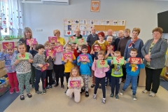 Na tle ściany stoi grupa przedszkolaków w kolorowych strojach. Trzymają w rękach wykonane przez siebie ozdobne kartki - laurki. Po obu ich stronach stoją nauczycielki. Z tyłu za dziećmi stoją osoby starsze - 5 kobiet i 4 mężczyzn.