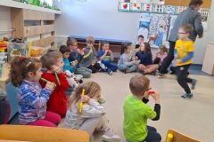 Grupa dzieci przedszkolnych siedzi w półkolu i gra na kolorowych fujarkach. Jeden z chłopców, ubrany w żółto - niebieską bluzę skacze w środku na jednej nodze.  Z boku  widoczna od tyłu przechodząca nauczycielka.