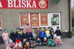 12 dzieci stoi w półkolu, 8 kuca. Za nimi stoi pani. W tle białe drzwi i 3 okna, na których widnieje znak apteki i napis APTEKA. U samej góry szyld- na czerwonym tle biały napis BLISKA.