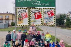 Część dzieci stoi, część kuca na zielonej trawie. Wśród nich stoi pani w jasnej kurtce. Za nimi duża, zielona tablica- logo sklepu Cezar. Poniżej reklama 3 produktów: napoju Tymbark, płynu do mycia naczyń Ludwik, ryżu Albaris. Obok każdego na czerwonym tle białymi cyframi wypisana cena.