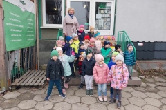 Grupa dzieci w kolorowych kurtkach I czapkach stoi przed sklepem na schodach. Z obydwu stron są zielone barierki. Przy nich na samej górze stoi pani w jasnej kurtce. Obok nich skrzynki z sadzonkami kwiatów. Za ich plecami szklane drzwi i 2 okna. W jednym widnieje stojak z powieszonymi torebkami nasion.