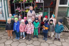 Dzieci stoją w grupce na szarych płytach. Wśród nich pani. Za nimi regał, na którym ustawione są bukiety ze sztucznych, kolorowych kwiatów, a także różne artykuły ogrodnicze.