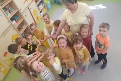 Sala przedszkolna. Grupa dzieci ubrana w odcieniach koloru żółtego stoi w zwartym kole Dzieci trzymają jedną ręką obręcz z żółtymi wstążkami. Z dziećmi w kole stoi dorosła osoba
